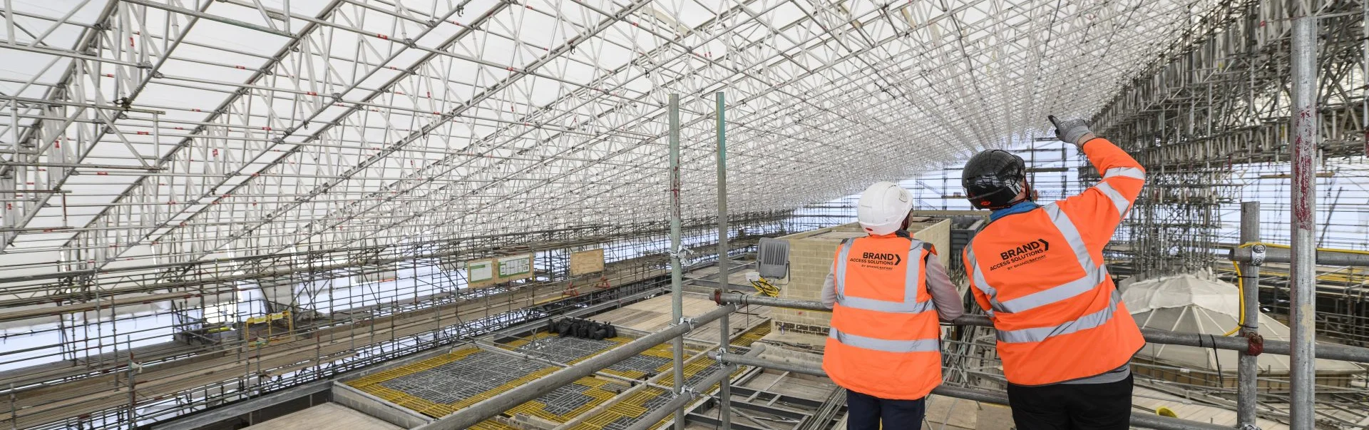 Two operatives in high-visibility jackets and helmets stand on a scaffold platform, looking toward a large temporary roof structure. A G-DECK system is visible to the left, used for safe access beneath the roof.